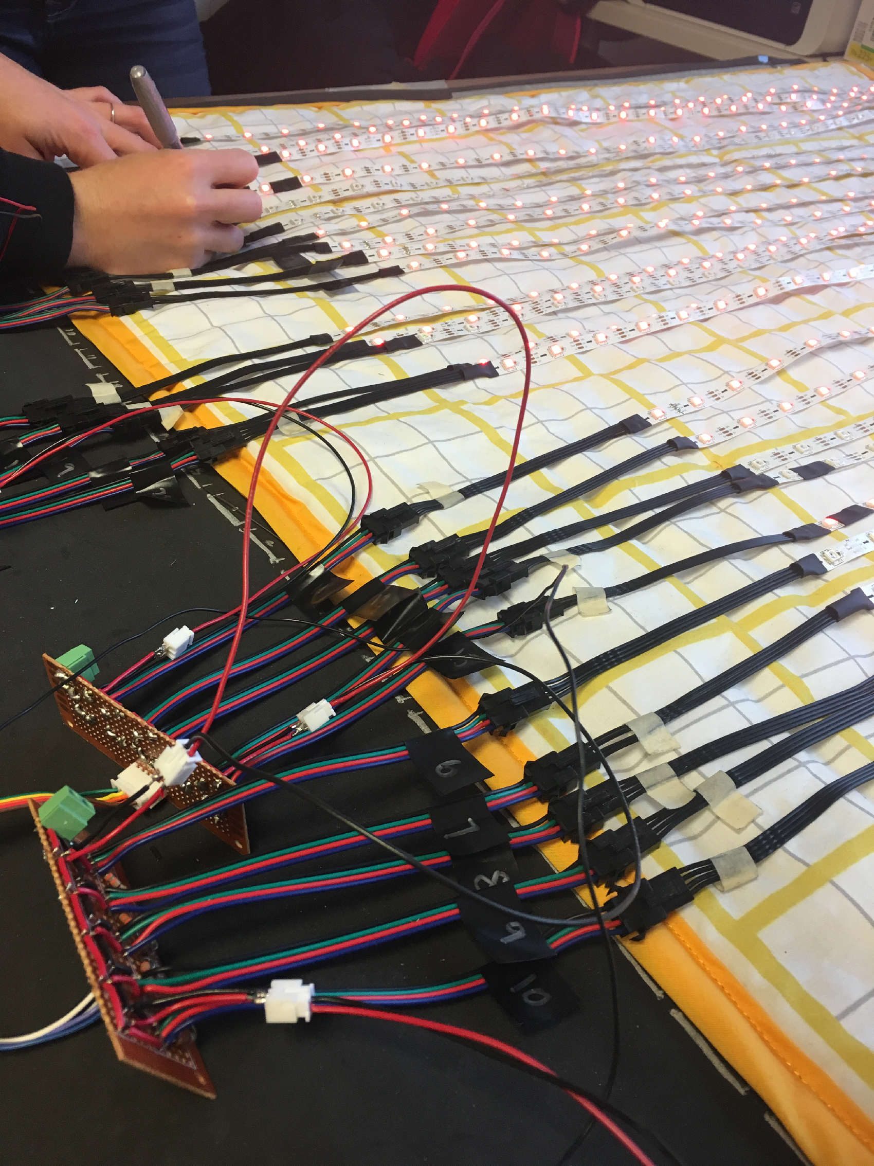 The third in a series of photos documenting the layers of the Floor Chart. Here, the LED strips are illuminated and arranged in a row on top of the quilt topper, and a pair of hands in the top left corner works with the cords.